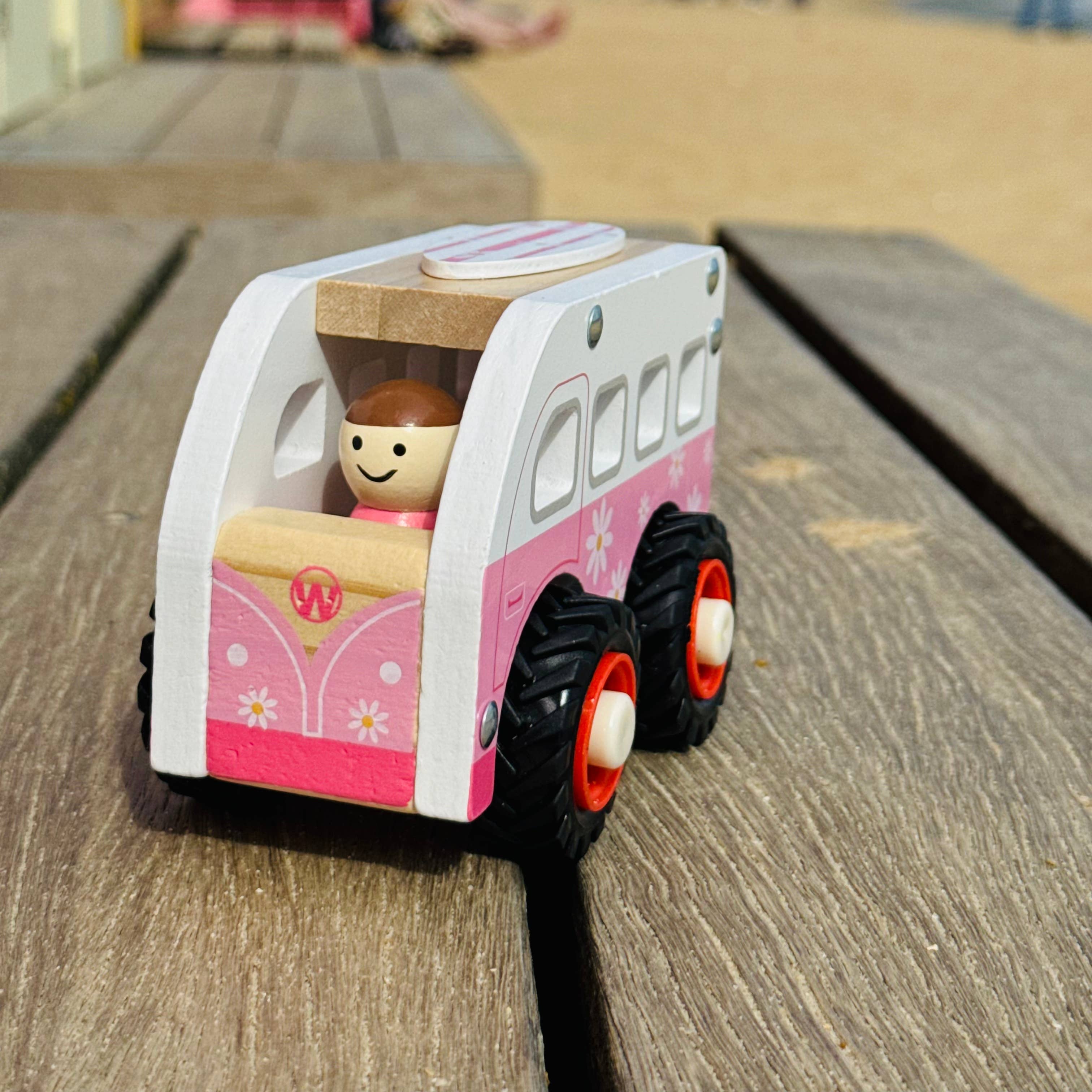 Toy camper on a wooden surface with a blurred background of people and a slide.