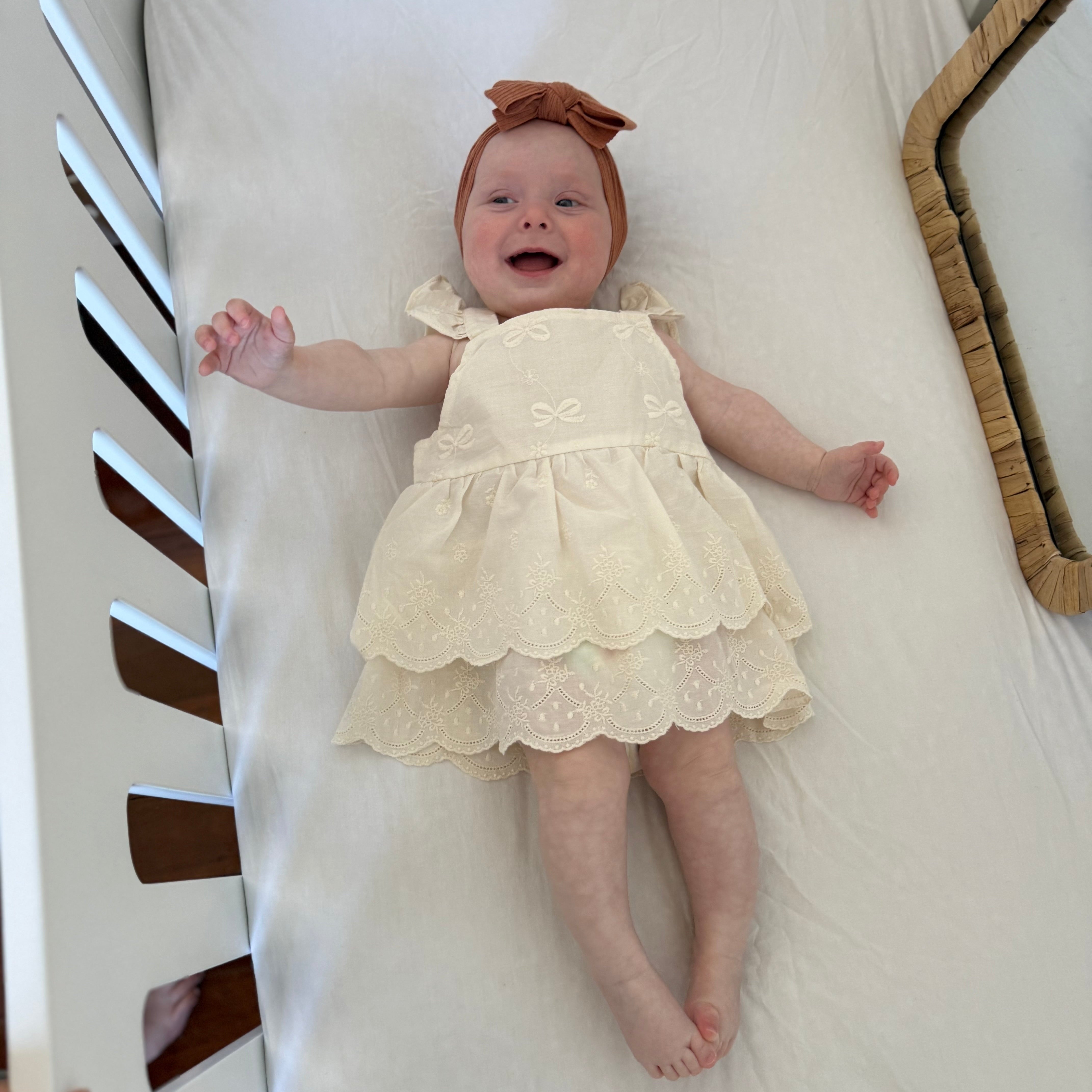 Baby in a white dress lying on a white blanket in a crib.