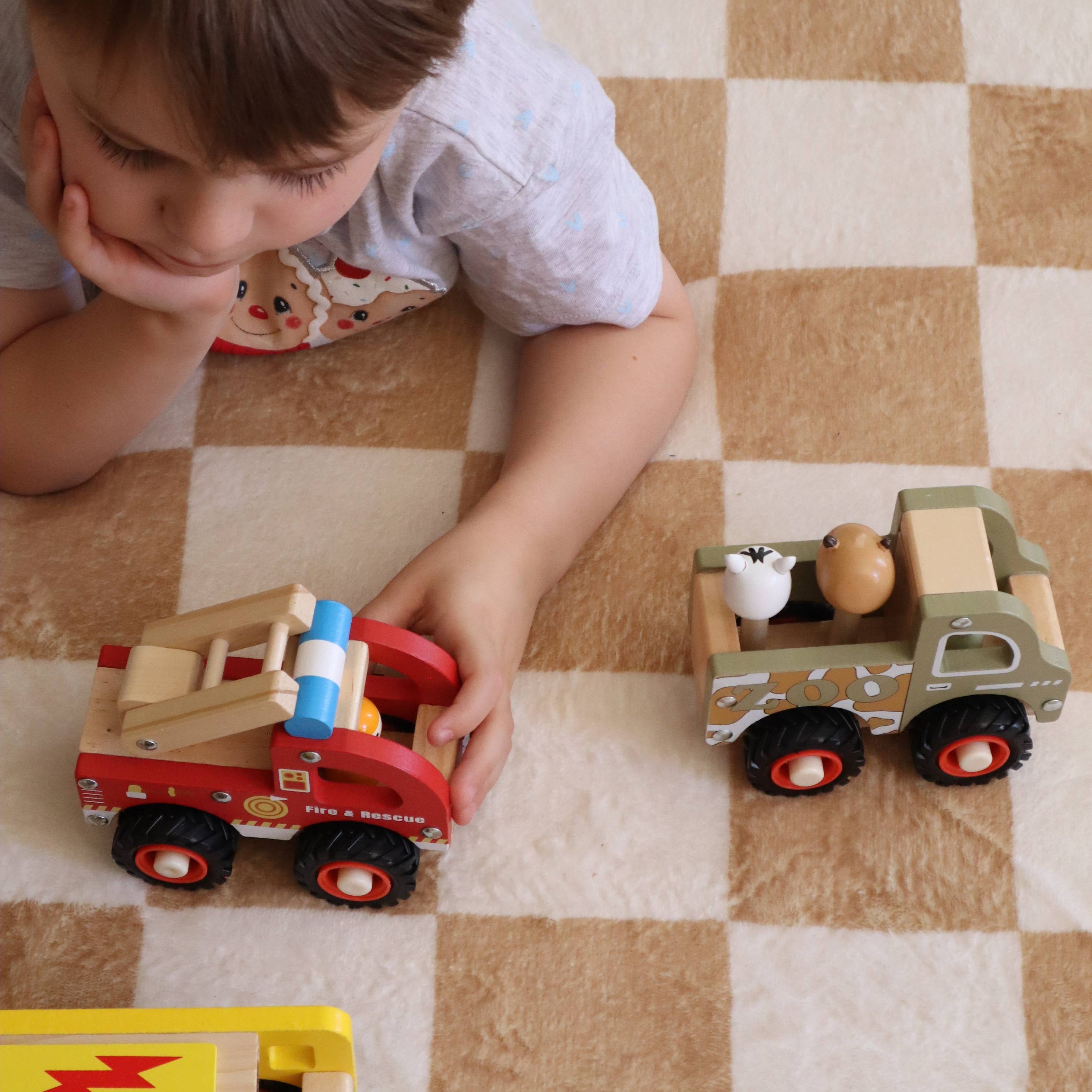 A young child lies on a beige and white checkered rug, playing with a set of wooden toy trucks, including a red fire engine and a green dump truck.
