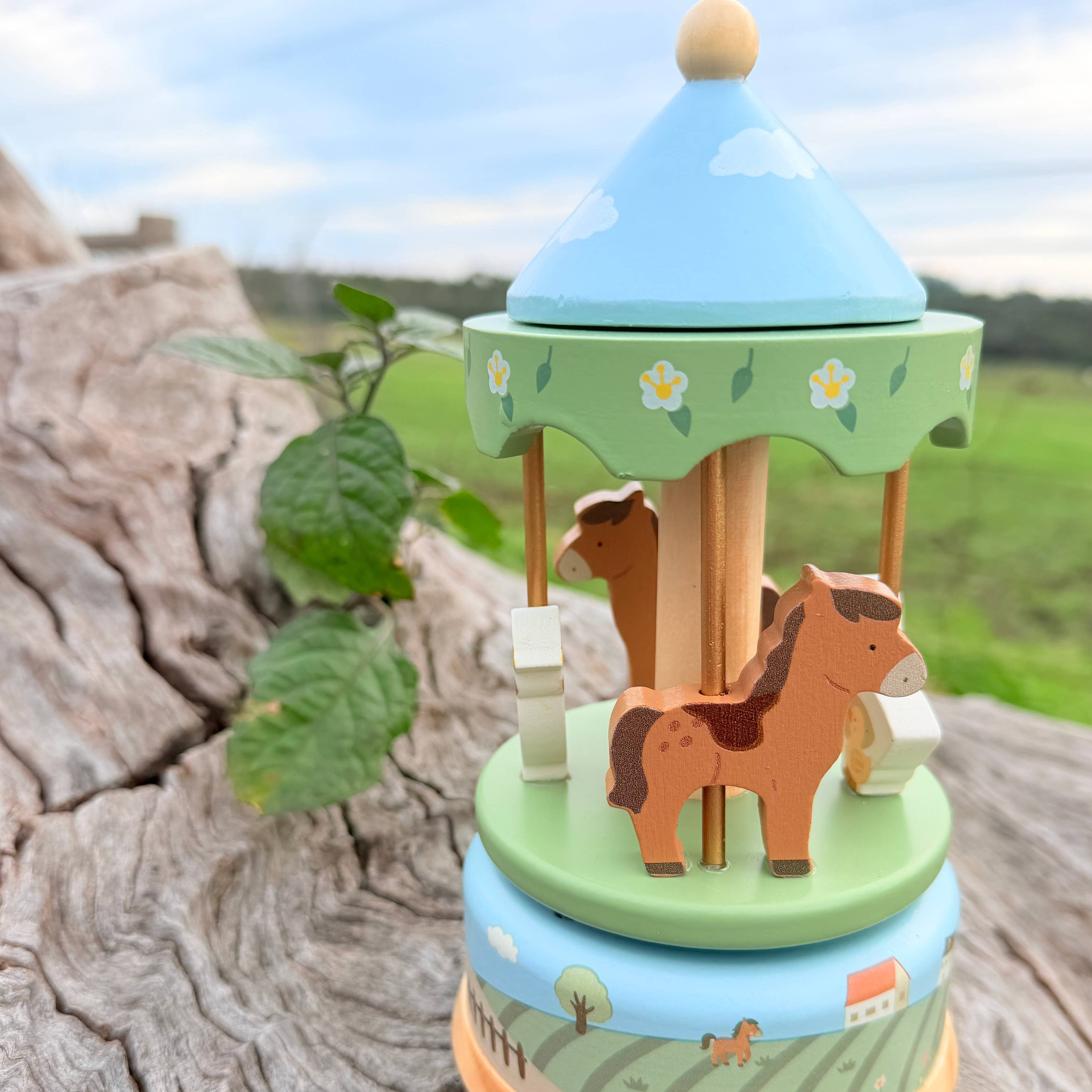 A small wooden carousel music box with a light blue top, green canopy, and brown horse figure, placed on a weathered log outdoors with a blurred field and sky in the background.