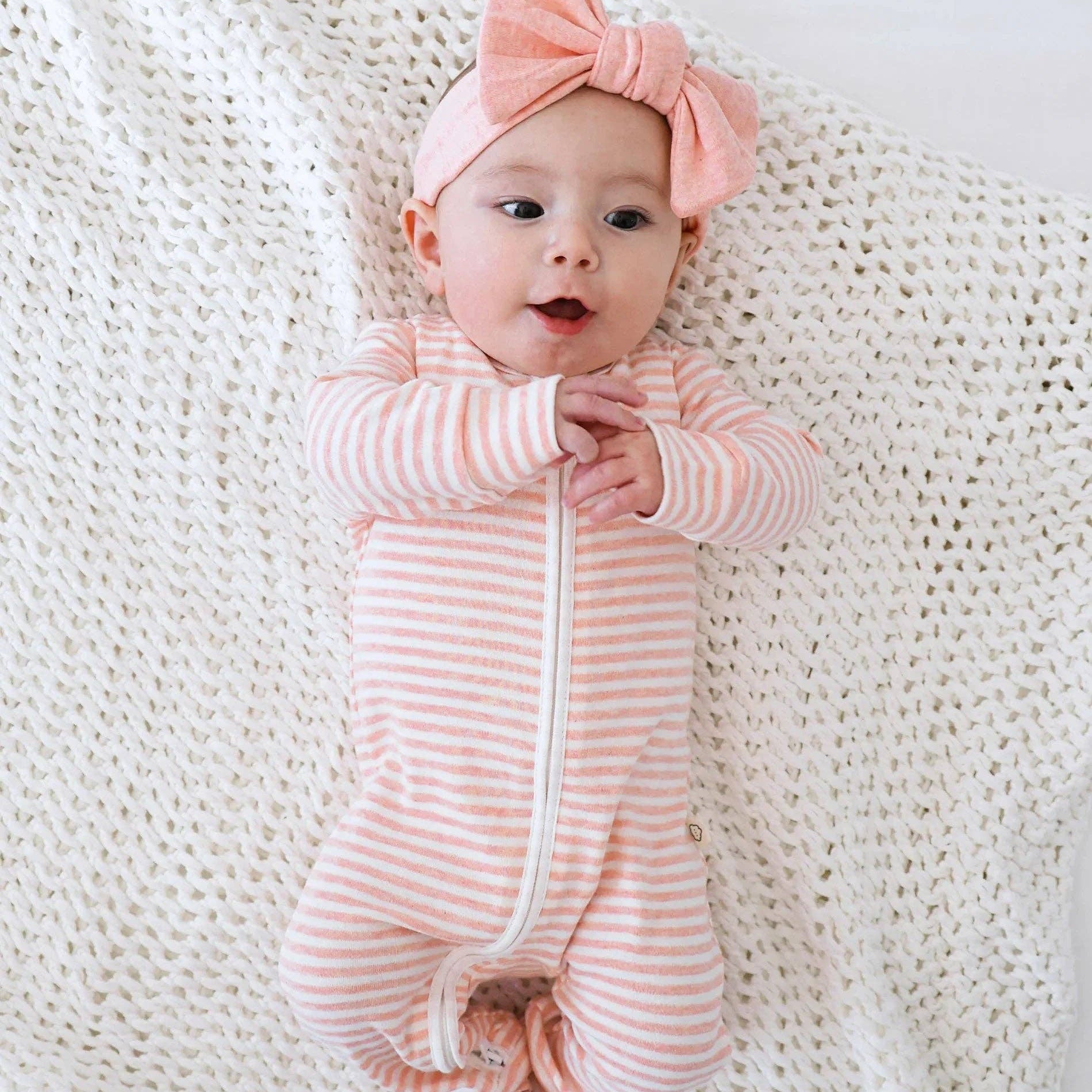 Baby in a pink and white striped outfit with a matching headband on a white textured blanket.