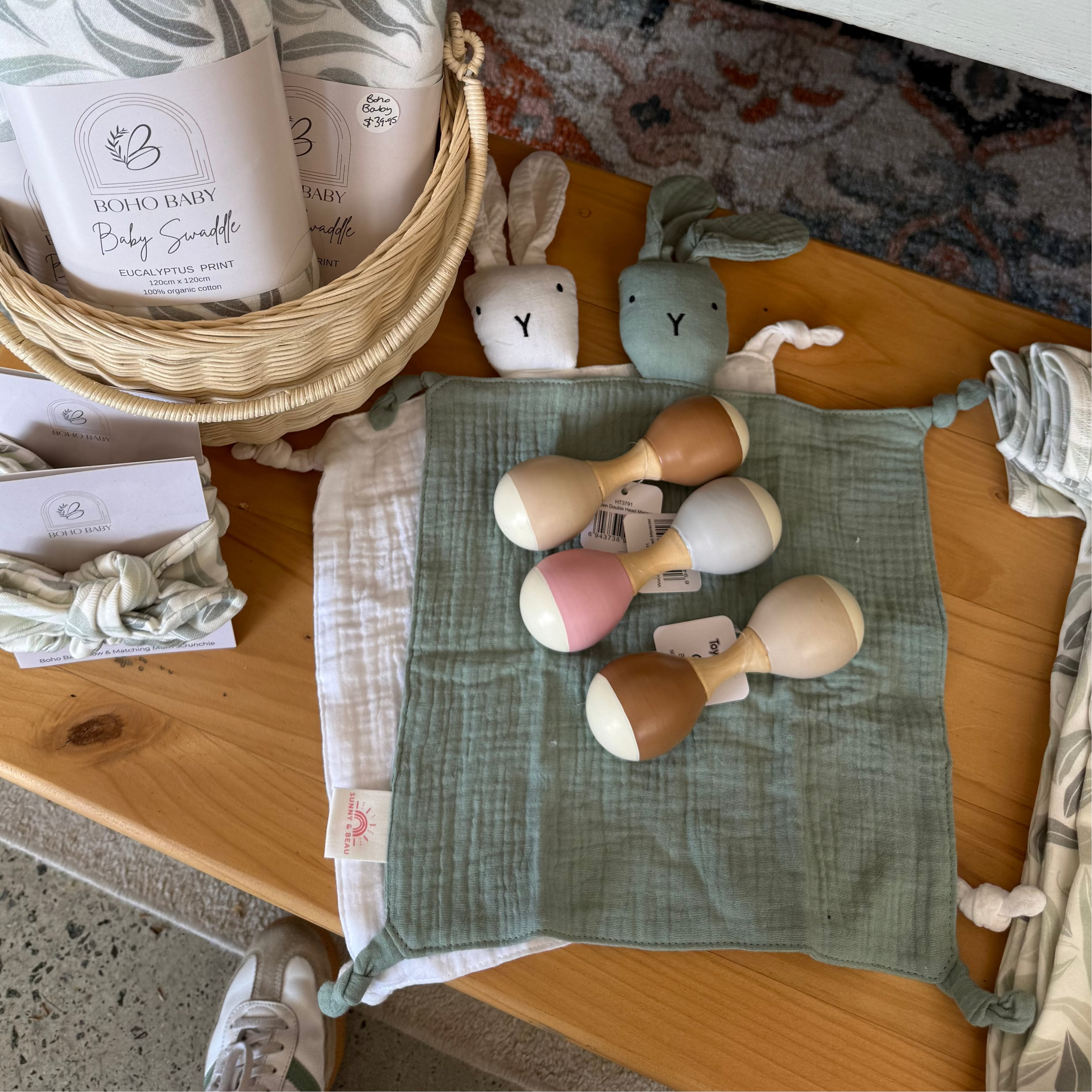 A baby gift set arranged on a wooden bench, including a woven basket with patterned baby swaddle blankets, wooden baby teething toys, a sage green muslin cloth, and small cards. The brand name 'Soothe & Soft' is visible on the blanket packaging.