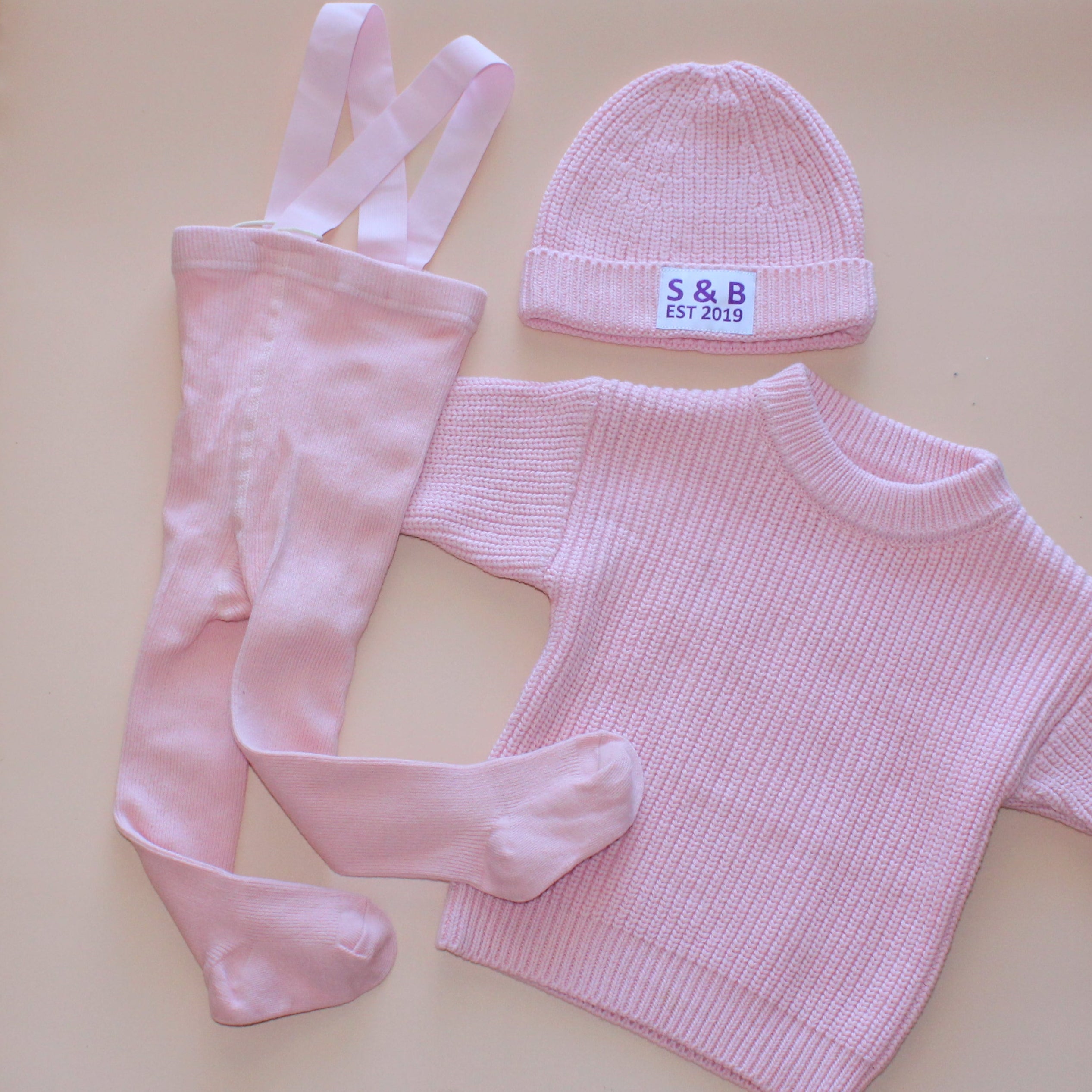 Pink baby outfit with hat, pants, and sweater on a beige surface.