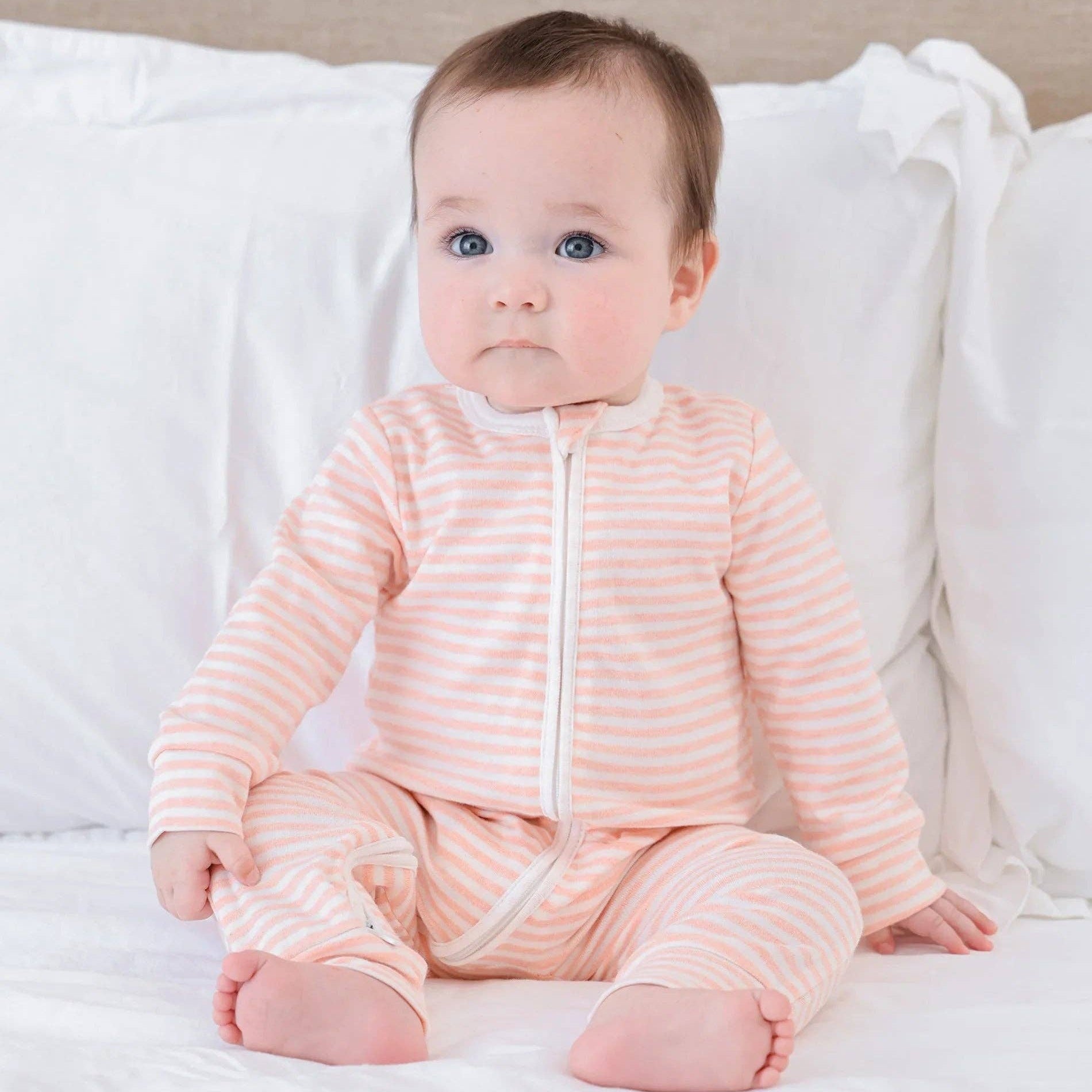 Baby wearing a pink striped onesie sitting on a white bed.