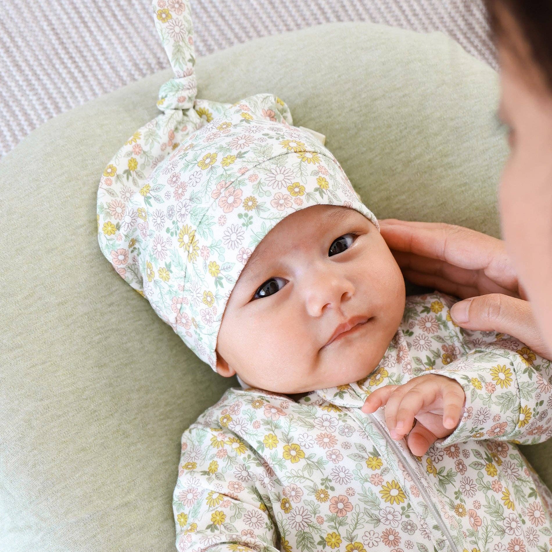 Baby in floral outfit and hat being held on a green cushion