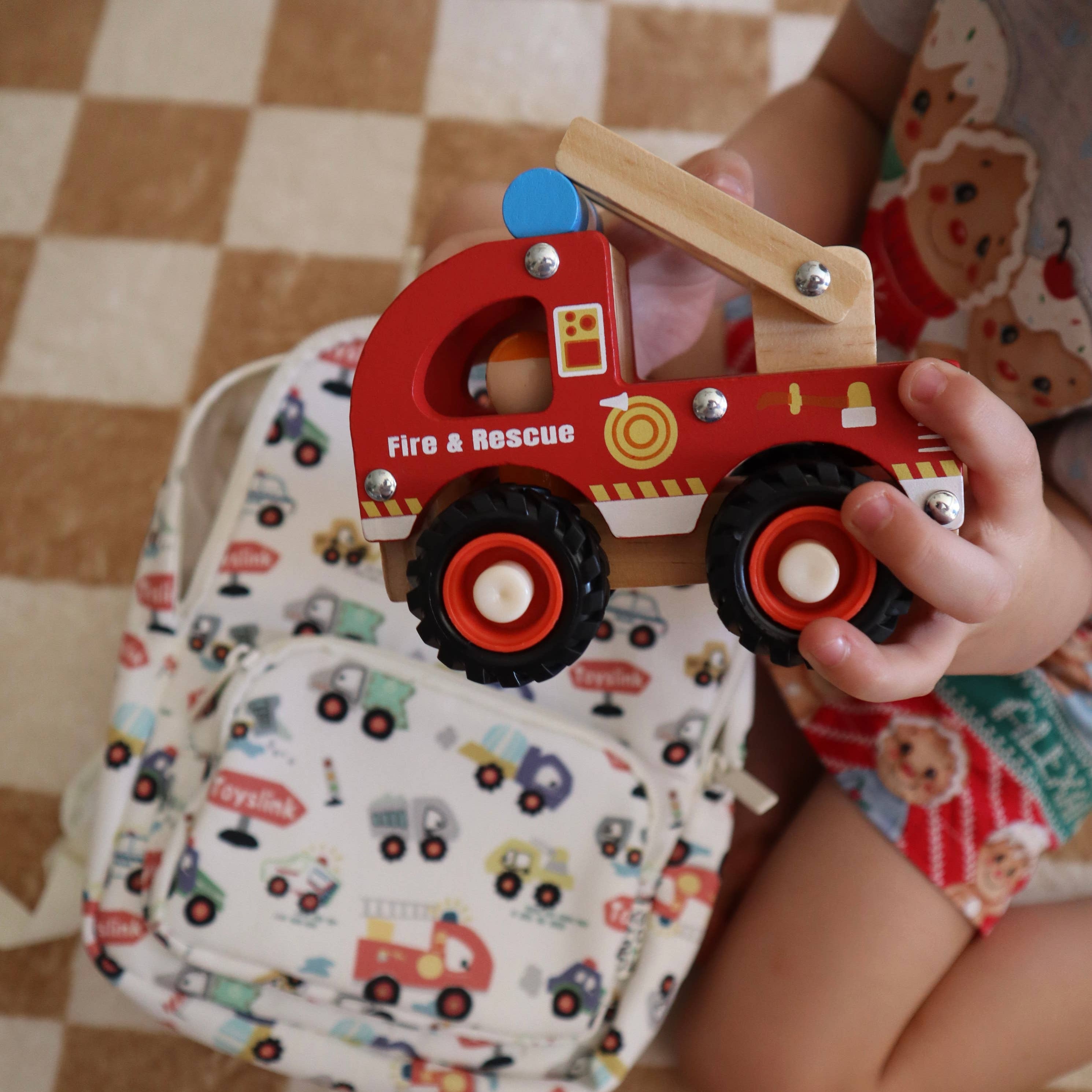 A child holds a red wooden fire truck toy with black wheels and a light wood crane arm, sitting on a brown and beige checkered floor next to a white backpack with a colorful fire truck pattern.