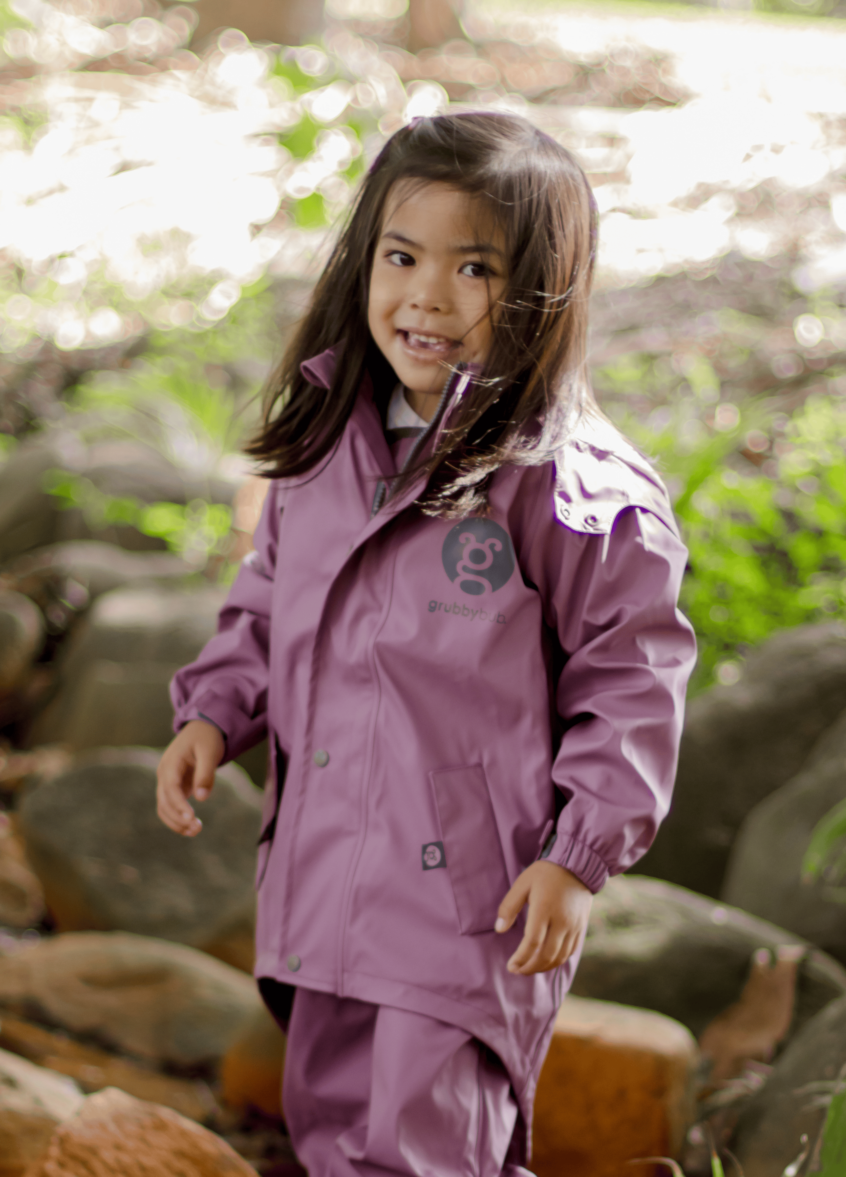 Young girl wearing a purple raincoat standing outdoors with greenery in the background