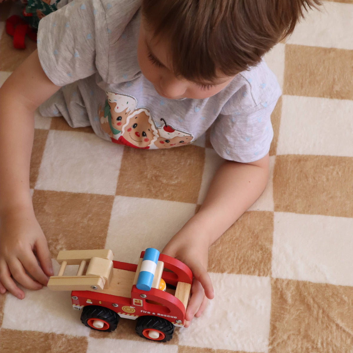 A young child with brown hair wearing a light gray t-shirt lies on a beige and white checkered floor mat, playing with a wooden toy set consisting of a red fire truck and a yellow cargo trailer.