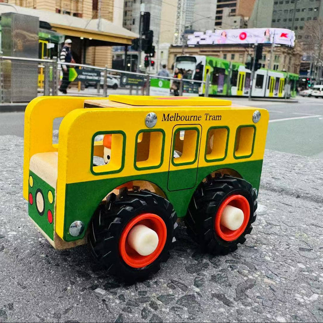 A wooden toy bus shaped like a Melbourne Tram, painted in the classic yellow and green color scheme, positioned on a city street with a blurred urban background.