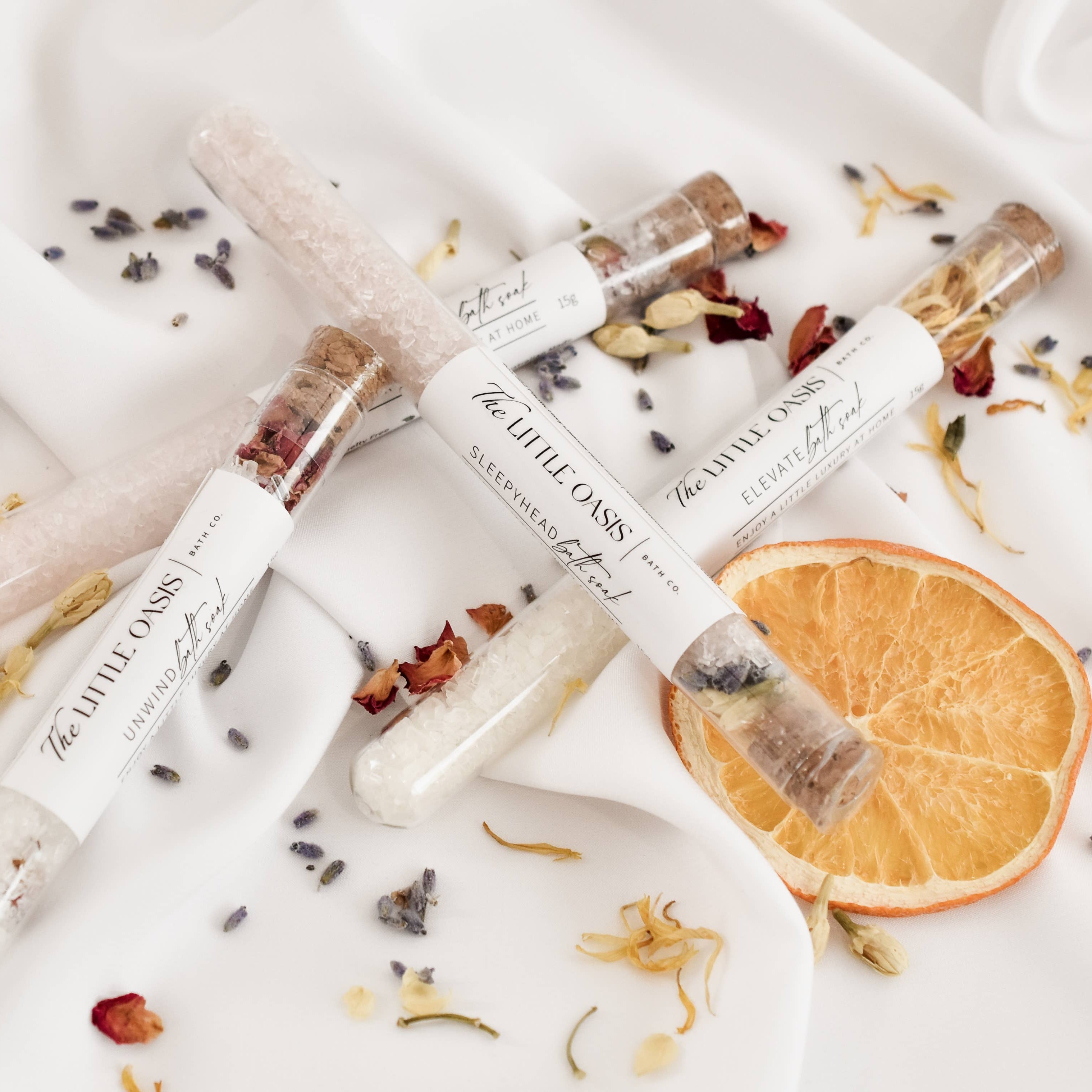 Three small glass vials with bath salts, herbs and flowers on a white surface with dried orange slices and flowers.