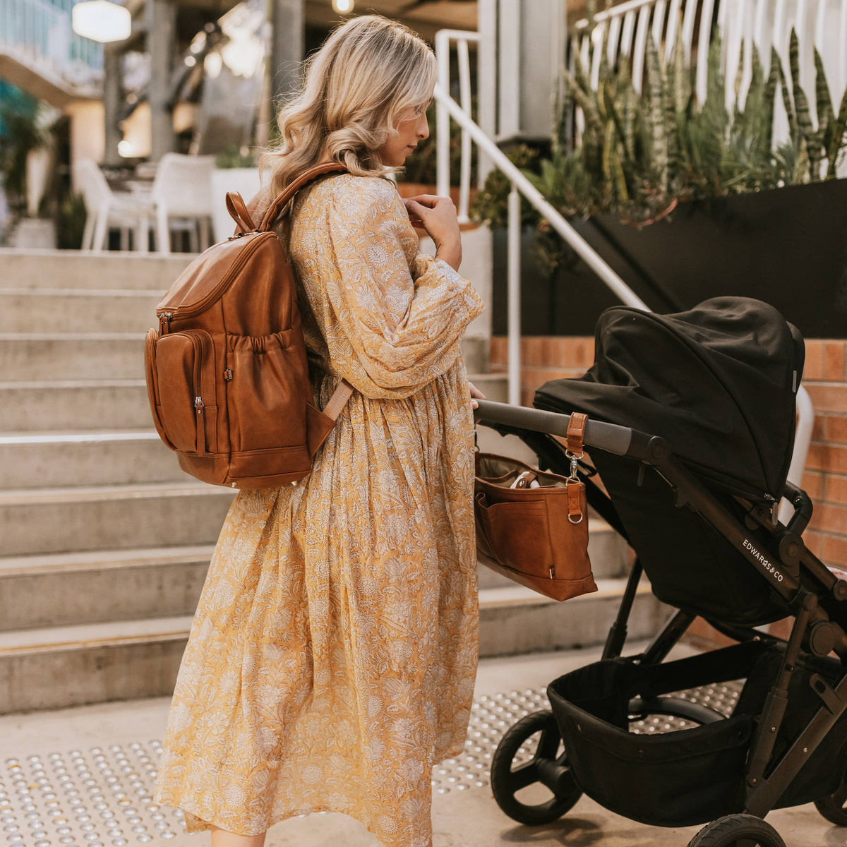 Woman in a yellow dress with a brown backpack and stroller, standing on a patio.