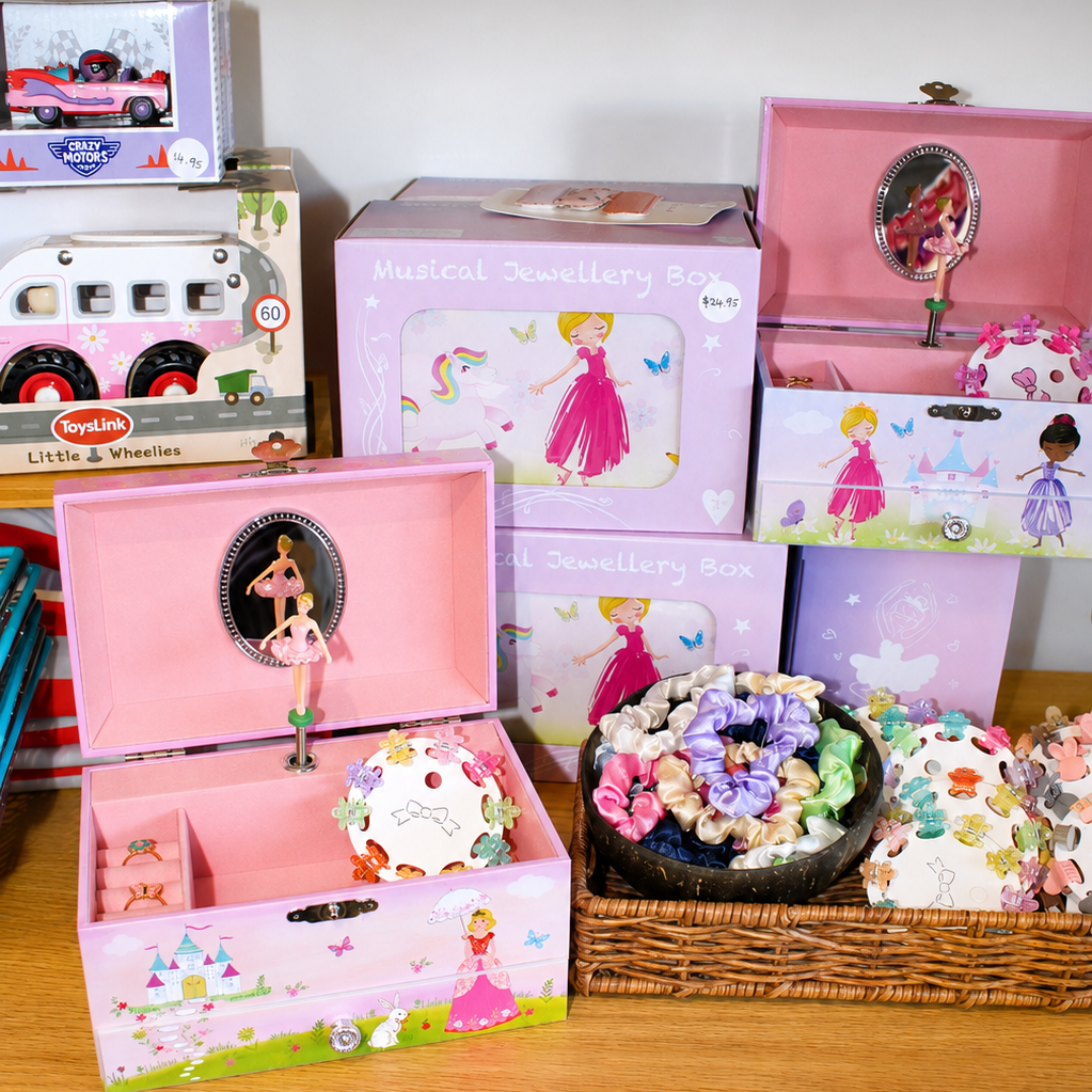 A collection of pink and pastel-themed musical toy boxes and decorative boxes displayed on a wooden shelf, including a pink music box with a fairy window, a pink music box with a doll, and several matching decorative boxes.