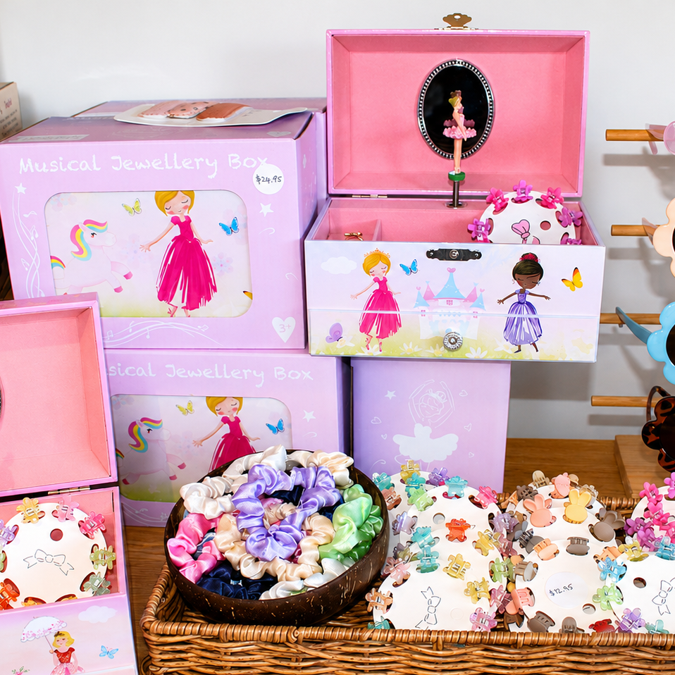 A collection of pink and purple children's jewellery boxes with ballerina illustrations, a wicker basket filled with jewelled hair bows, and a wooden display holding pink, peach, and blue flower-shaped sunglasses.