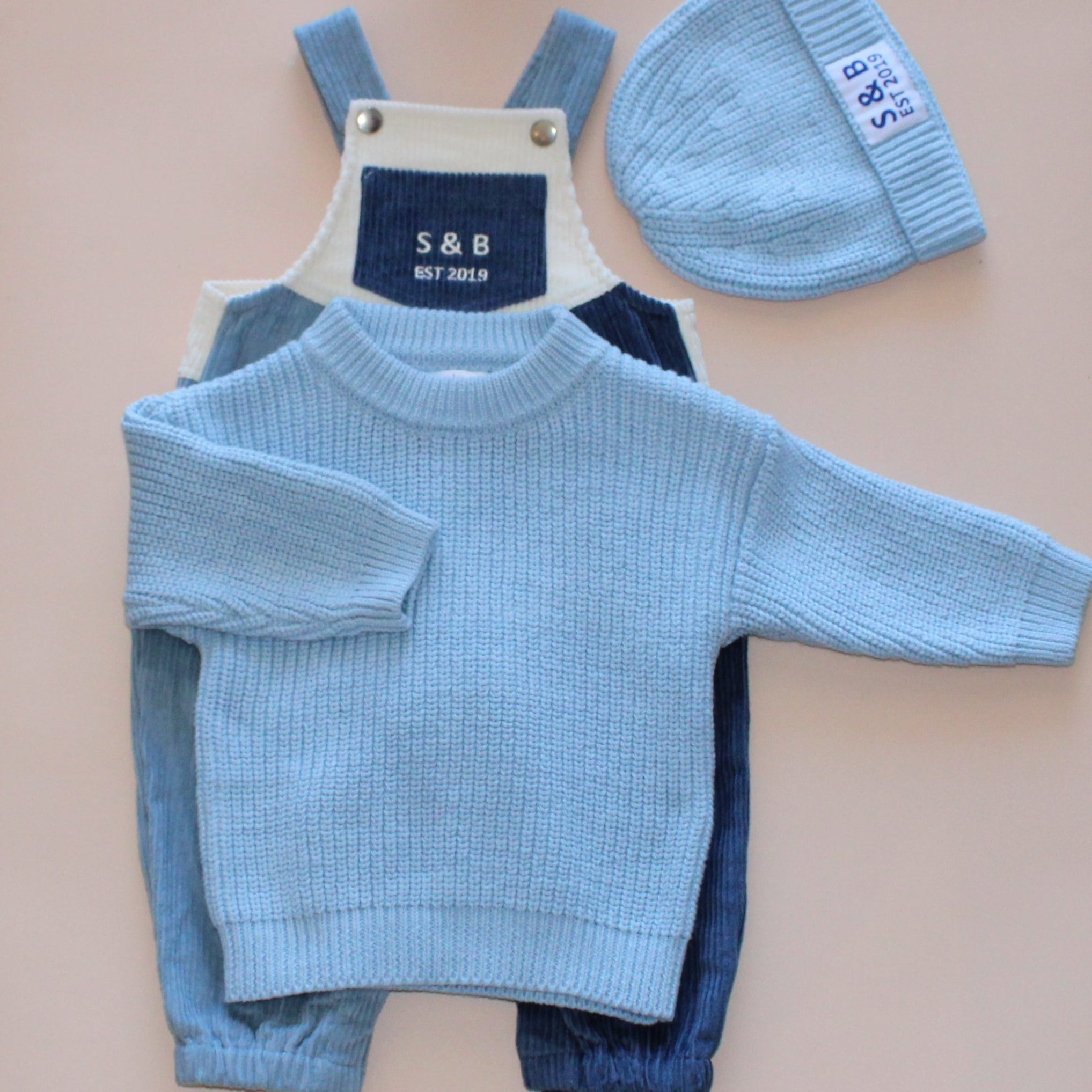 Baby outfit with blue sweater, pants, and hat on a pink sheet on a concrete floor.