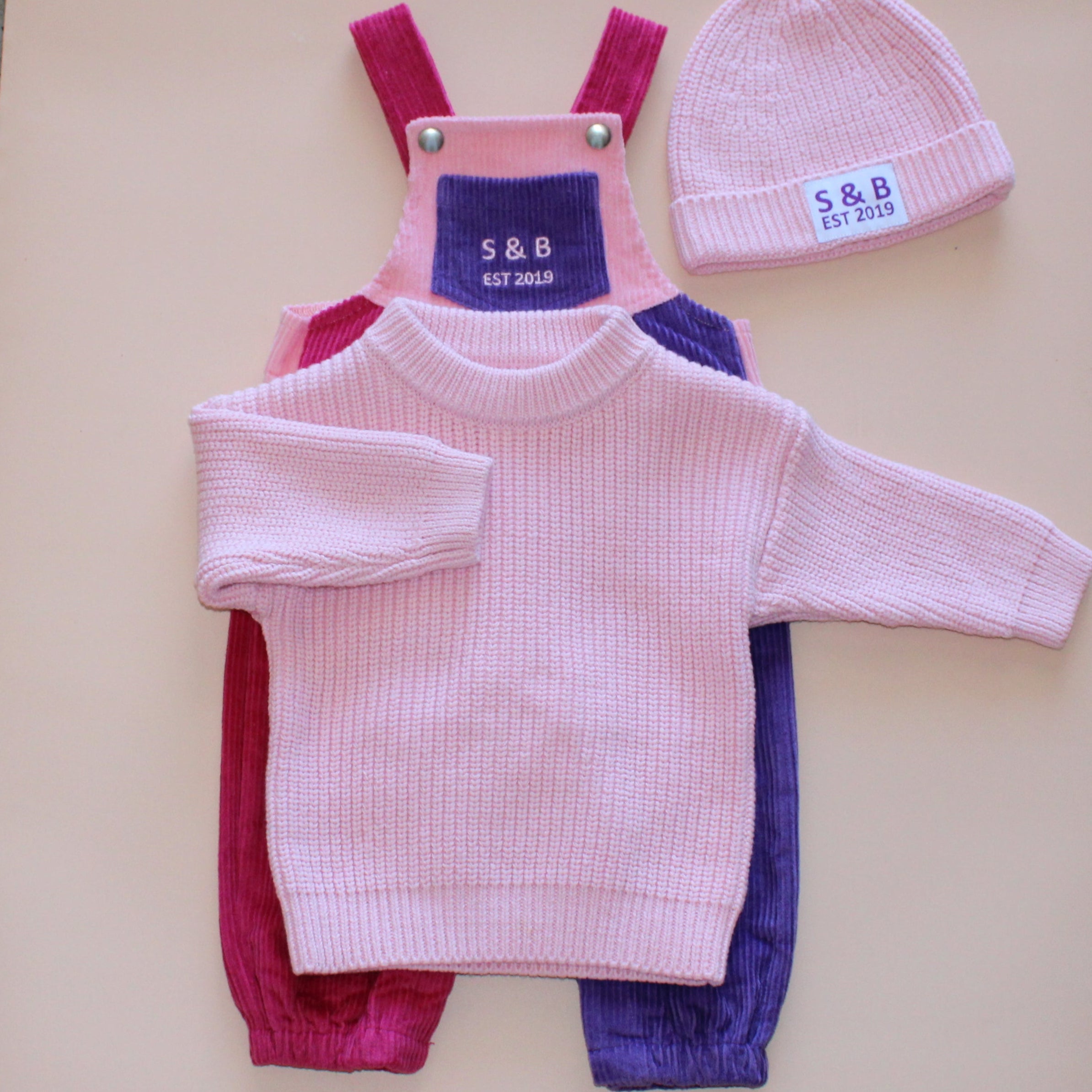 Children's pink outfit with overalls and hat on a beige surface, with a concrete floor background.