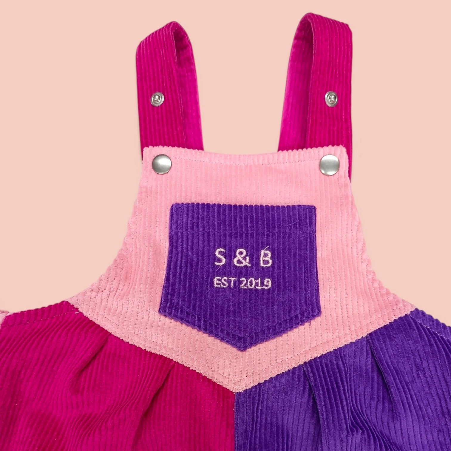 Pink and purple corduroy overalls with a pocket and initials 'S&B' on a beige background