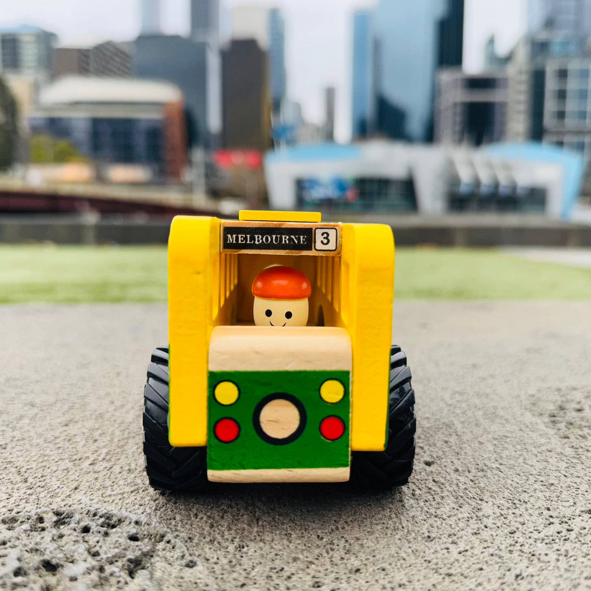 A small wooden toy bus with a yellow body, green front panel, black wheels, and a wooden driver figure, photographed outdoors on a paved surface with a blurred city skyline in the background.